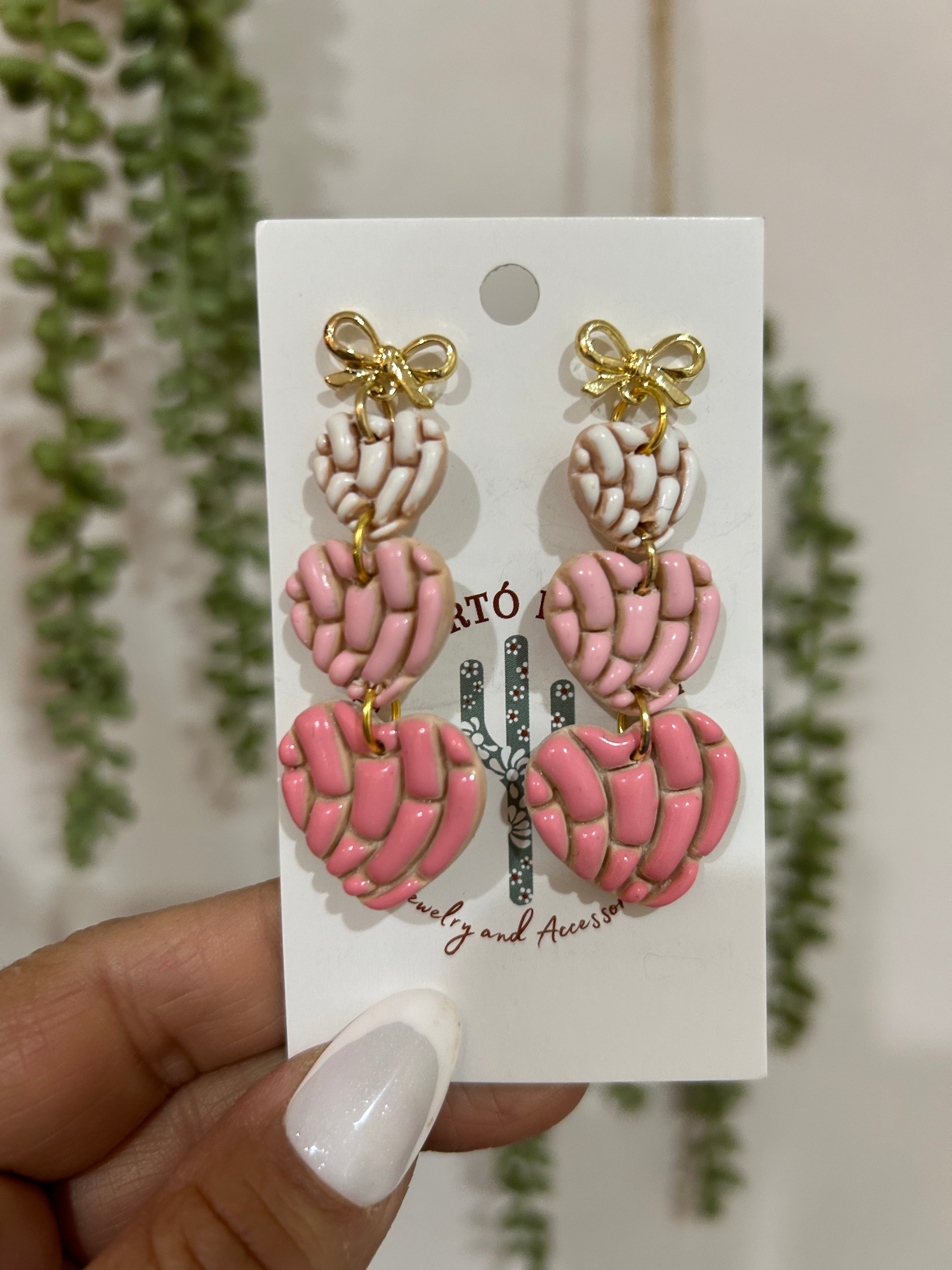 Set of pink and gold concha heart earrings on a backing card held by a hand with a blurred green plant background