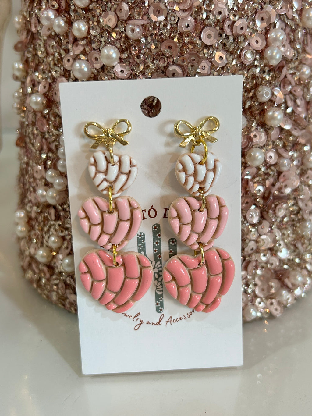 Pair of pink and white concha heart earrings with gold accents on a backing card against a glittery background.