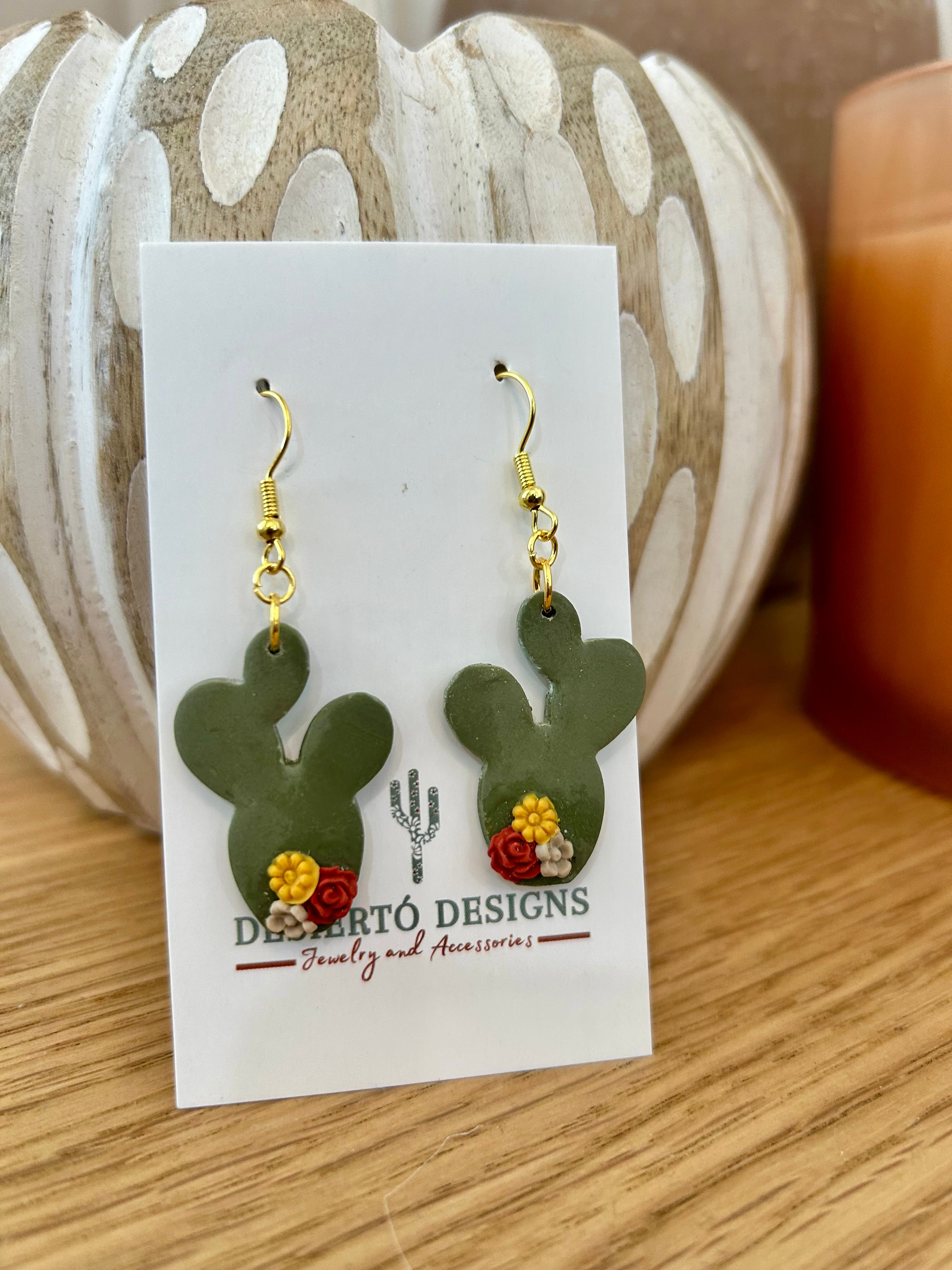 Cactus-shaped earrings with little flowers on a backing card against a wooden prop.