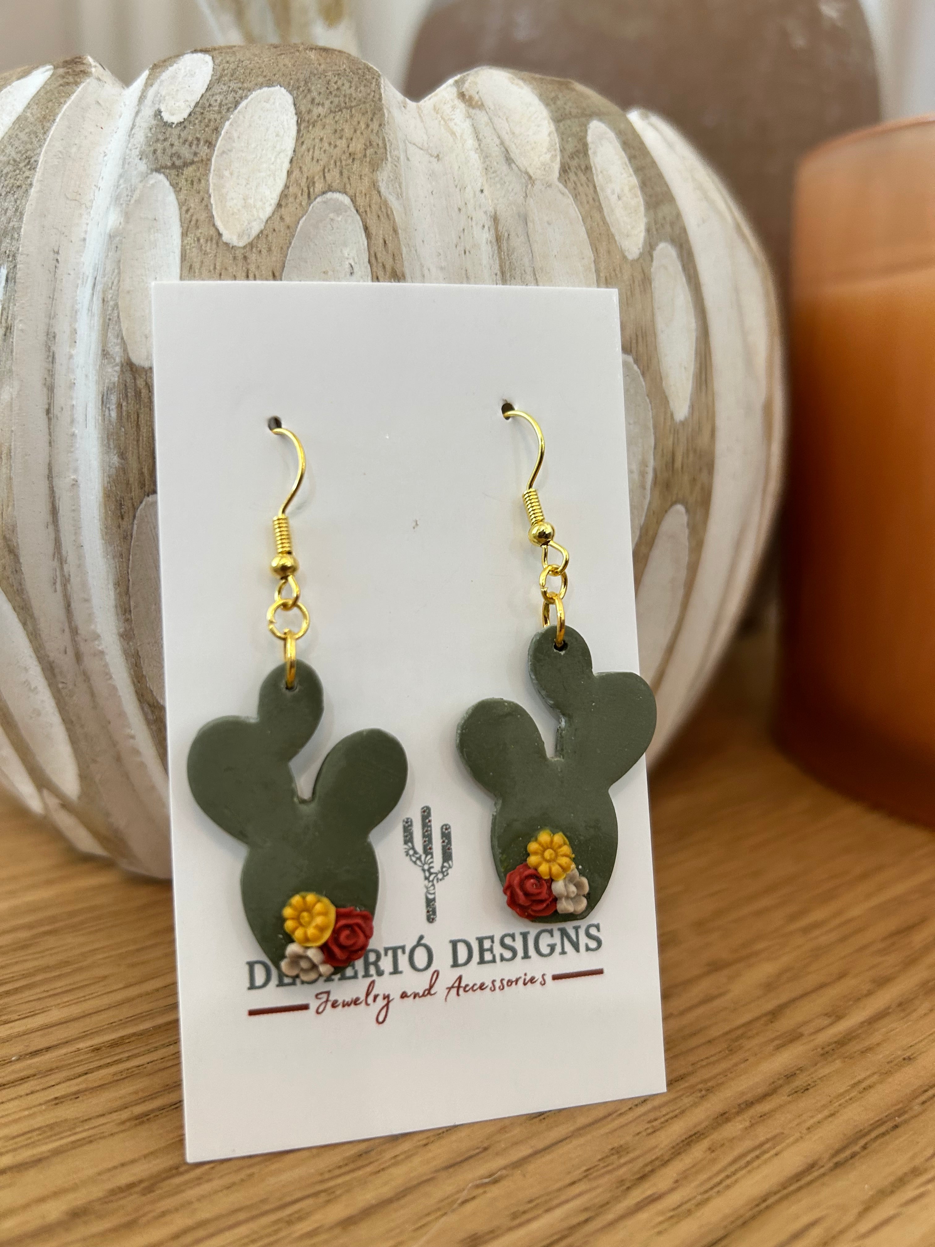 Cactus-shaped earrings with little flowers on a backing card against a wooden prop.