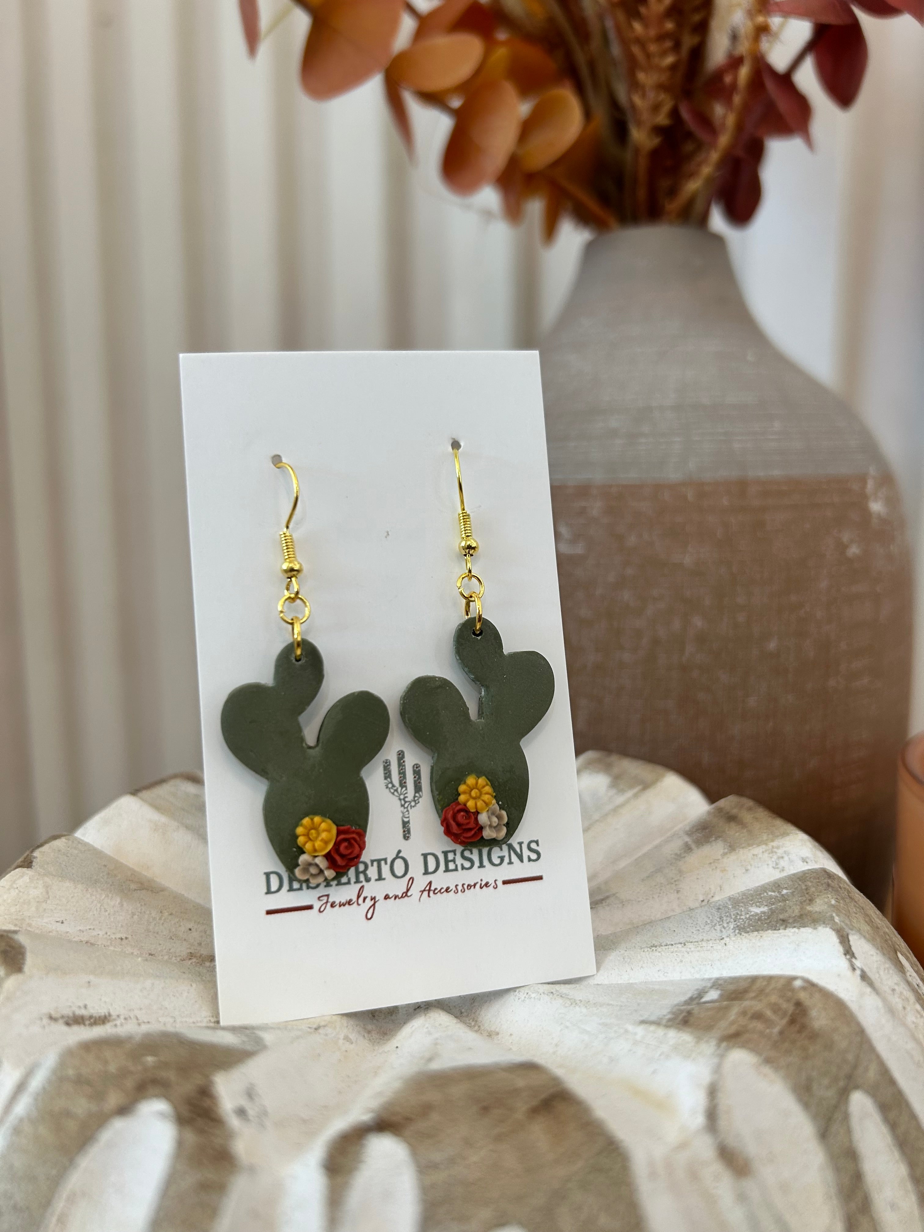 Cactus-shaped earrings with little flowers on a backing card on a wooden prop.