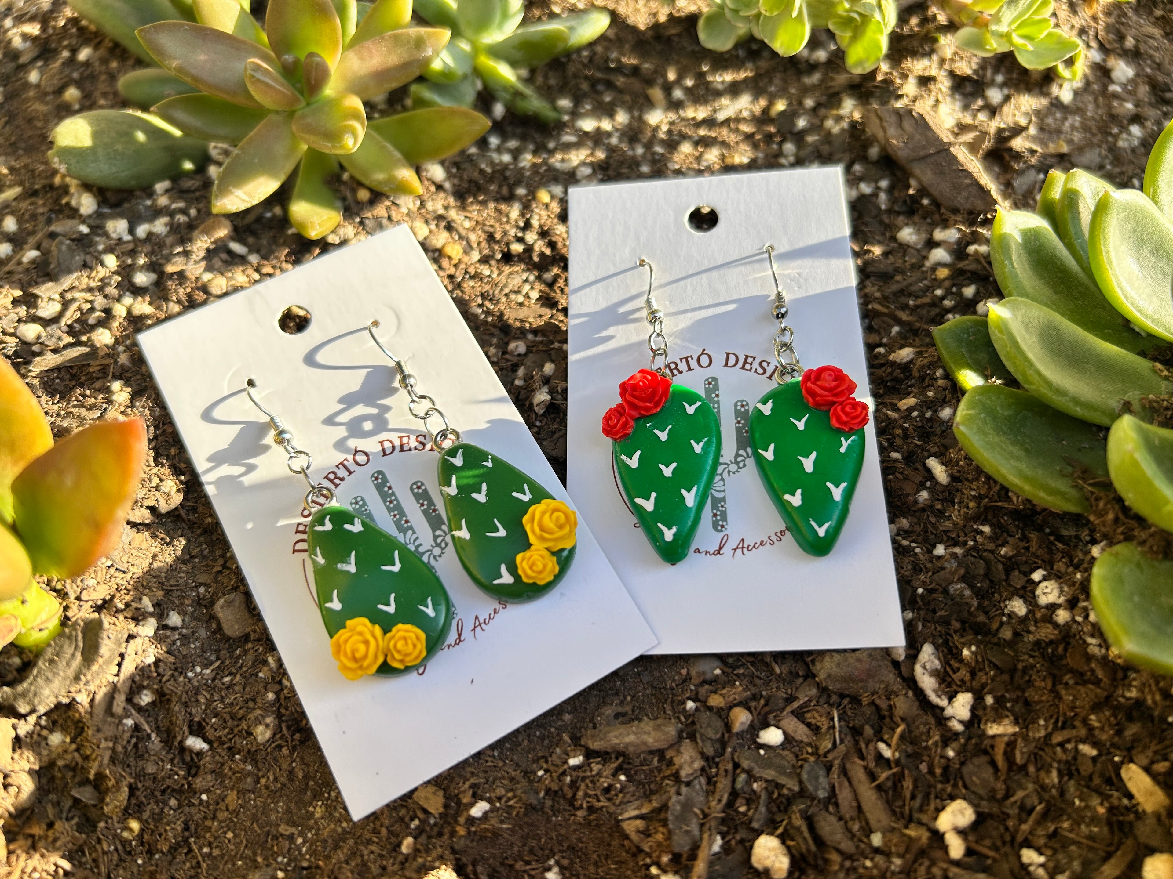 Cactus-shaped nopalito earrings with yellow or red rose flowers on backing cards surrounded by green succulents.