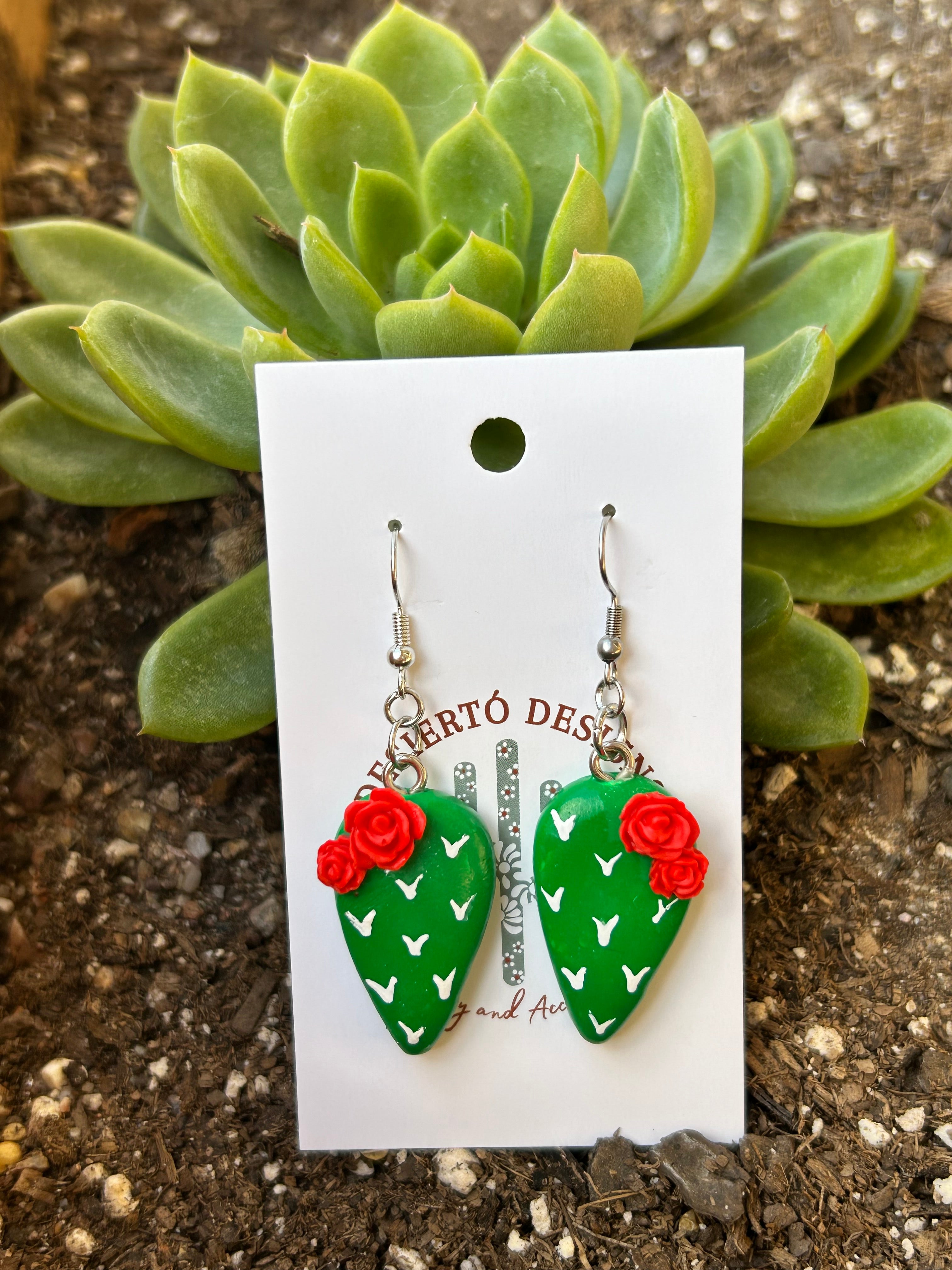 Cactus-shaped nopalito earrings with red rose flowers on a backing card against a green succulent.