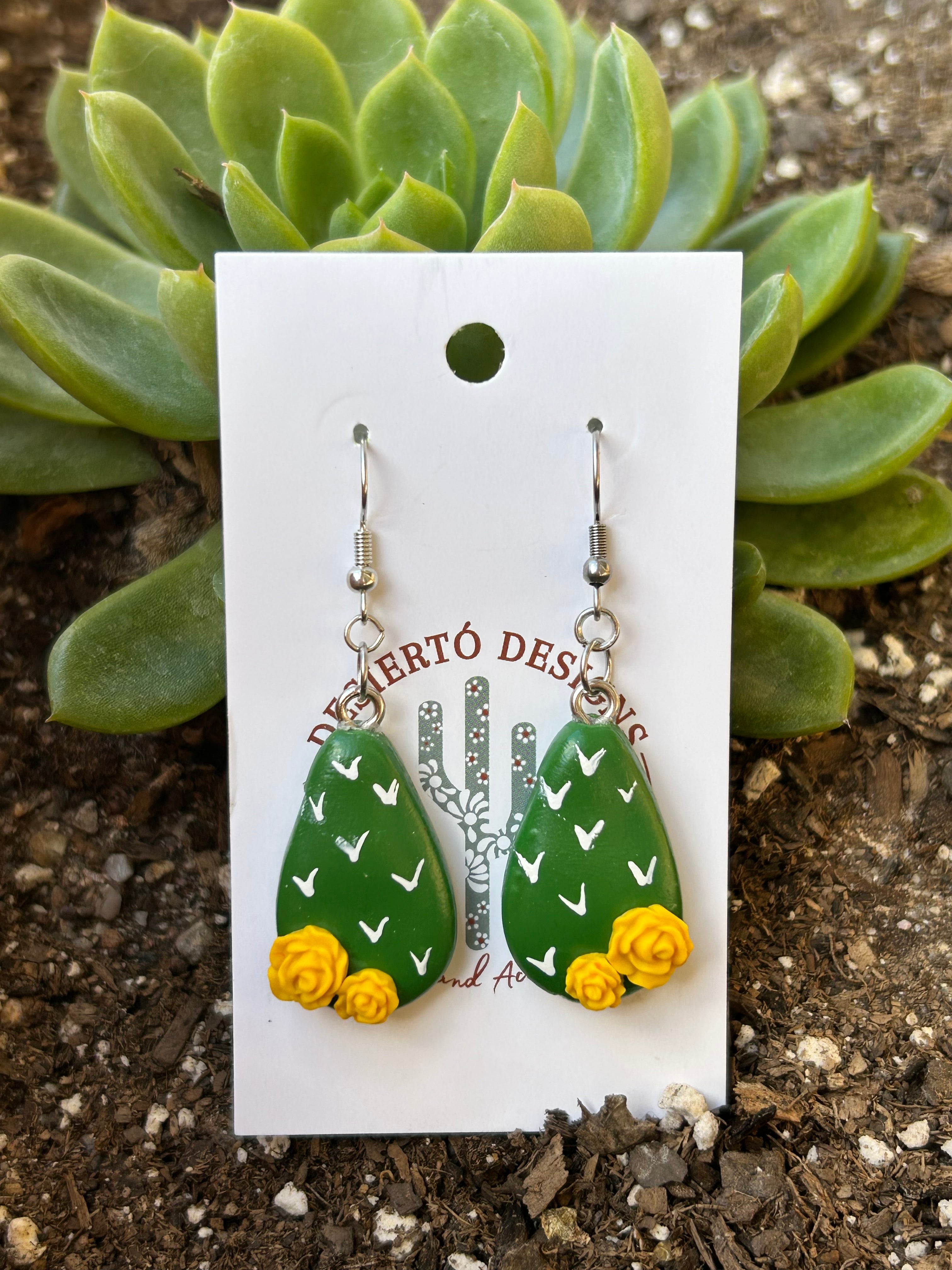 Cactus-shaped nopalito earrings with yellow rose flowers on a backing card against a green succulent.