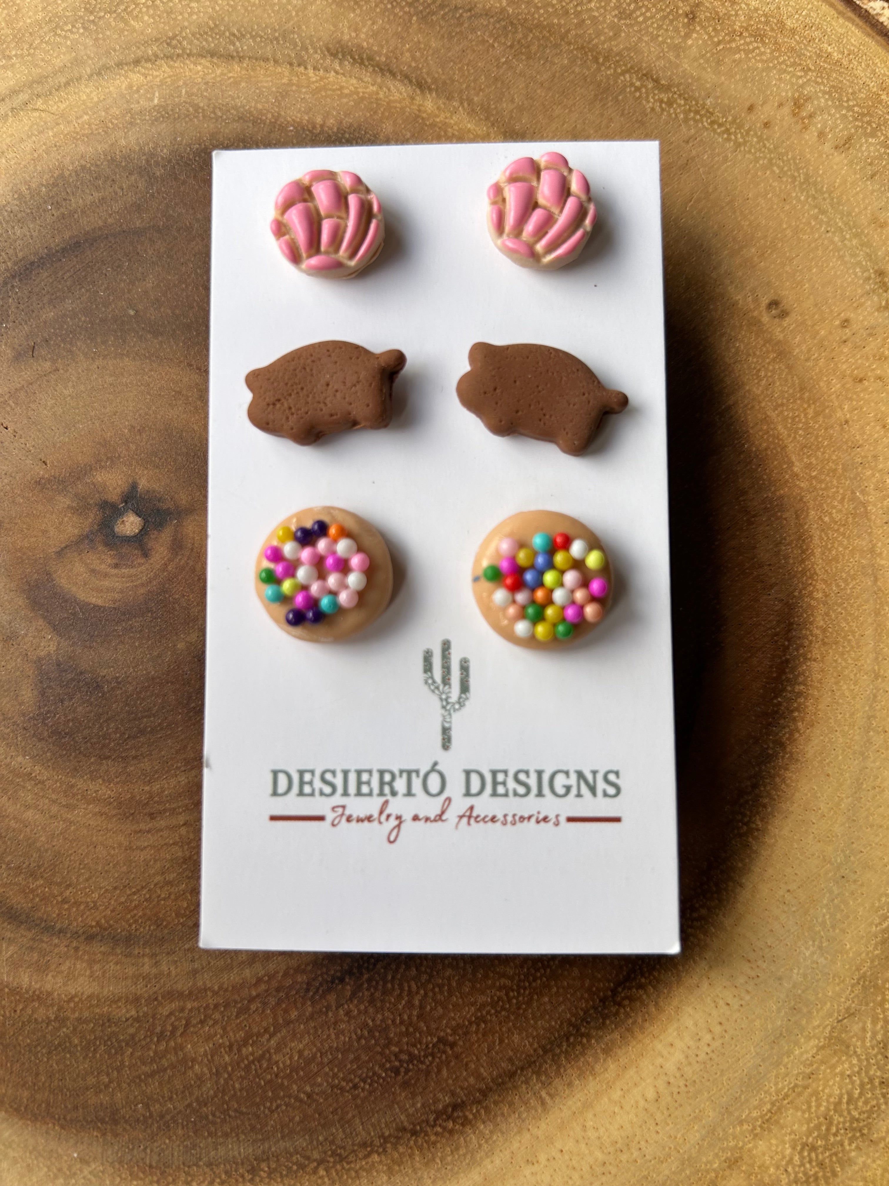 Three pairs of pan dulce stud earrings on a backing card against a wooden background.