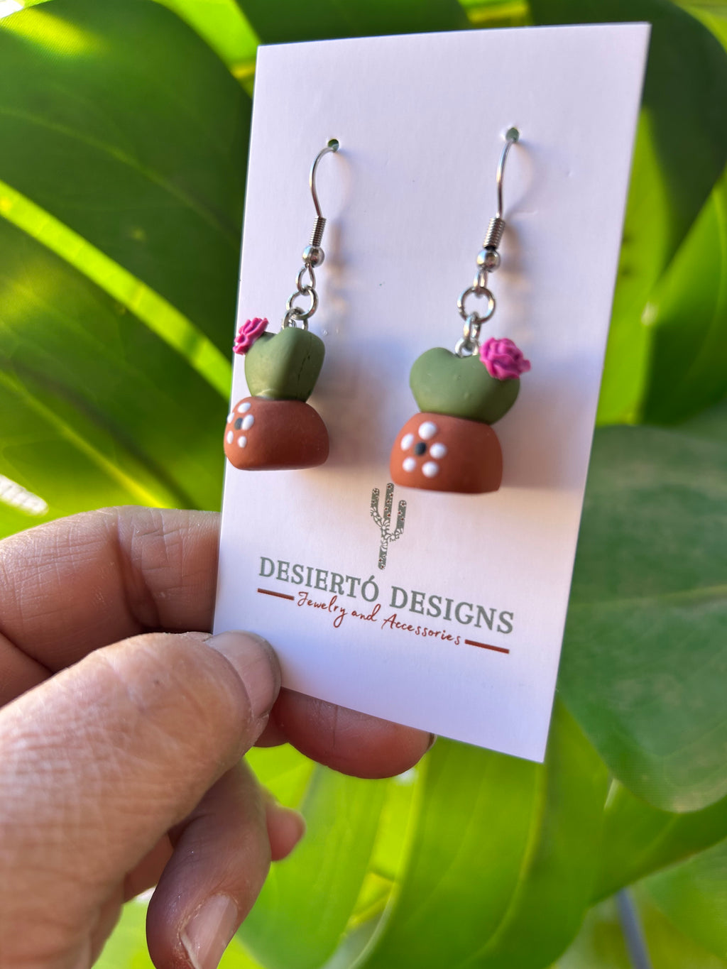 Potted Cactus Dangle earrings on a backing card with a green leafy background.