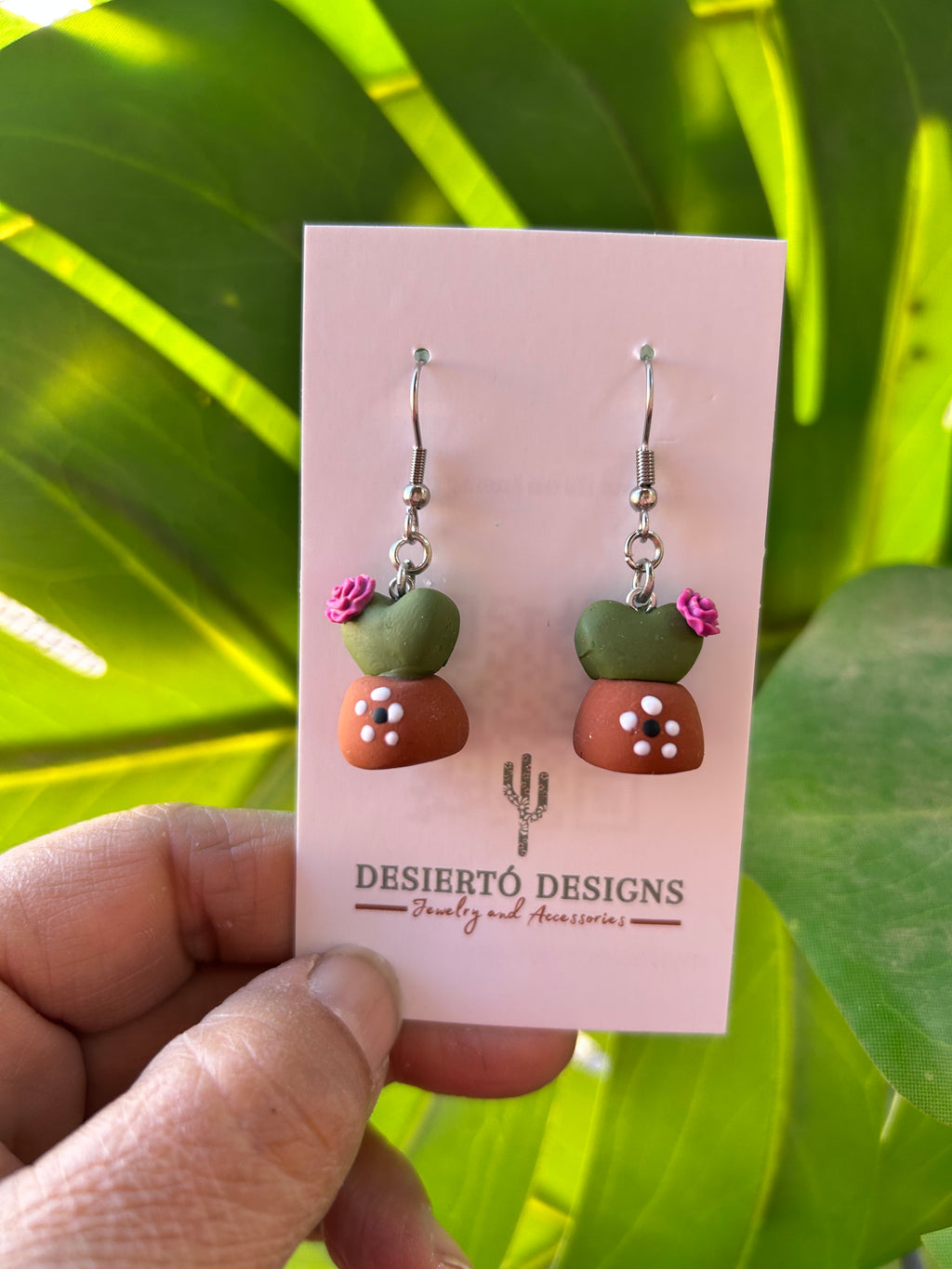 Potted Cactus Dangle earrings on a backing card with a green leafy background.