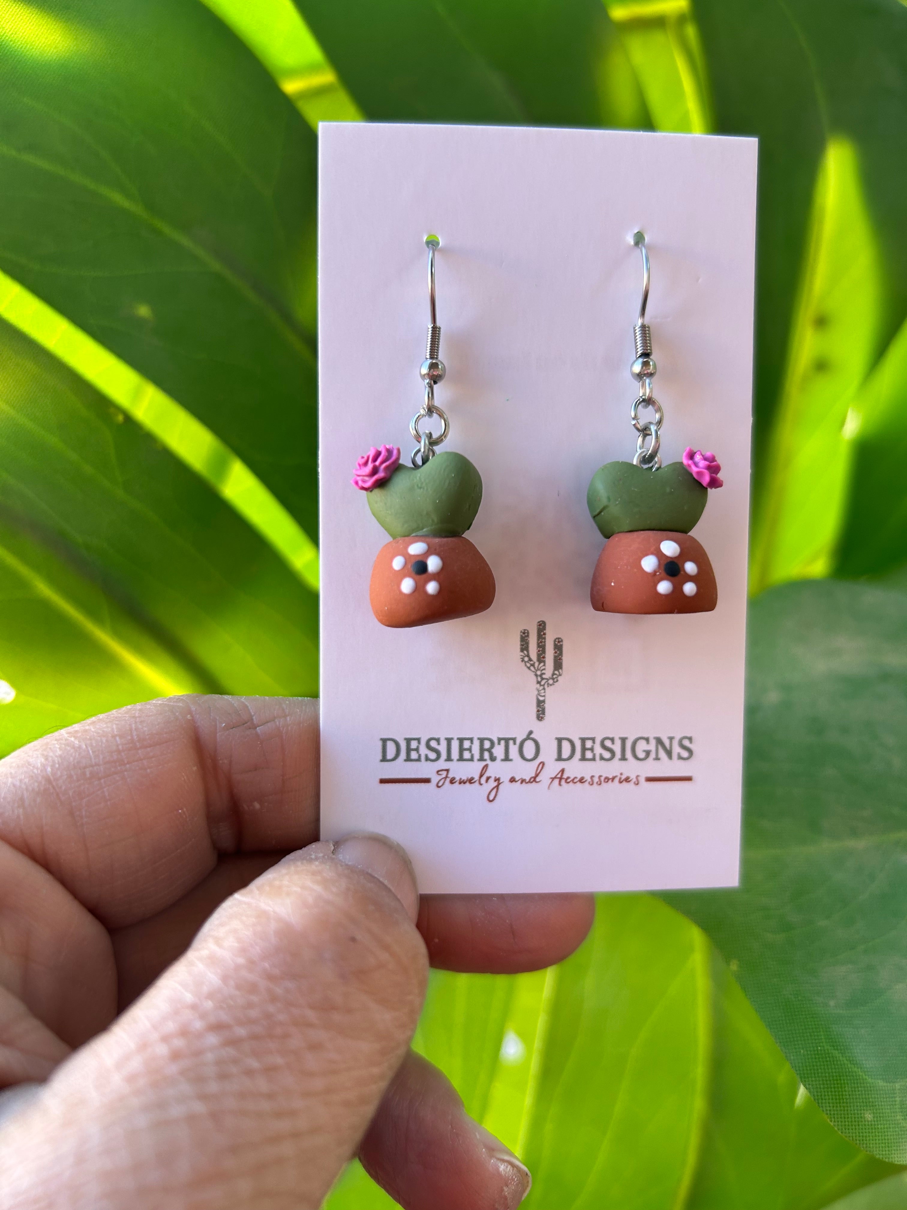 Potted Cactus Dangle earrings on a backing card with a green leafy background.